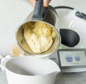 Eine Hand hält den Mixtopf des Thermomix®, darin ein Hefeteig, der in eine Schüssel fällt