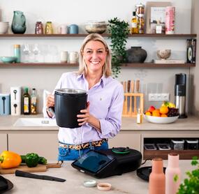 Blonde Frau hält den Mixtopf des Thermomix® in die Kamera und lächelt.