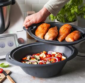 Zwei Hände halten den Einlegeboden des Varoma® mit Hähnchenbrustfilet darin. Im Varoma® liegt Gemüse, im Hintergrund steht der Thermomix®